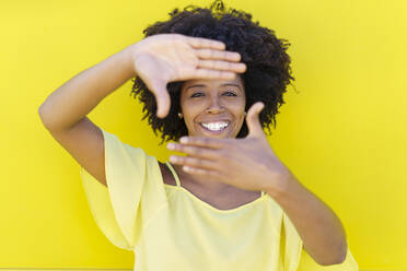 Happy young woman making frame with hands in front of yellow wall - JSMF02467