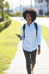 Student walking with mobile phone over footpath on sunny day - JSMF02478