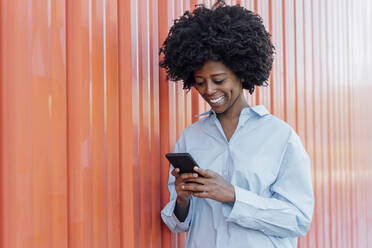 Smiling young woman using smart phone standing by orange wall - JSMF02481