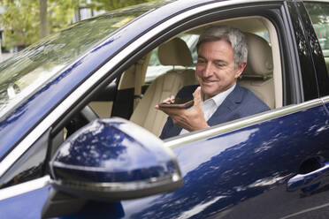 Smiling mature businessman sitting in car talking on speaker phone - SVKF00722
