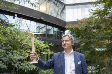 Smiling mature businessman with wind turbine model standing in office park - SVKF00733