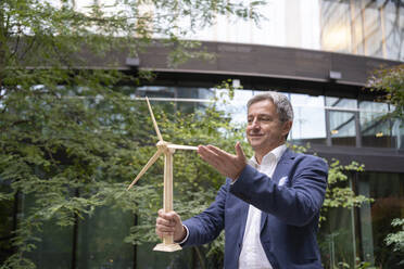 Mature businessman touching wooden wind turbine model at office park - SVKF00735