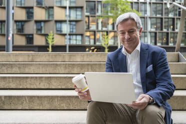 Lächelnder reifer Geschäftsmann sitzt mit Laptop und hält eine Einweg-Kaffeetasse auf einer Treppe - SVKF00740