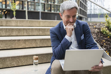 Mature businessman looking at laptop sitting on steps - SVKF00744