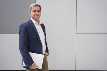 Confident mature businessman standing with laptop in front of gray wall - SVKF00746