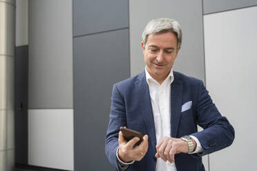 Älterer Geschäftsmann, der sein Handy hält und die Zeit auf seiner Armbanduhr vor einer grauen Wand überprüft - SVKF00750