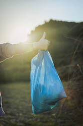 Junge Frau zeigt Daumen hoch Geste hält blauen Müllsack auf Sonnenuntergang - ACPF01528