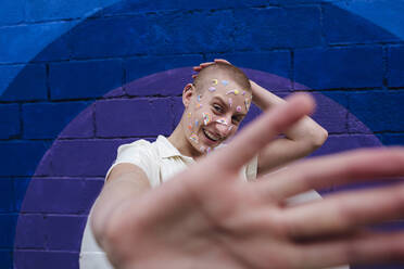 Happy non binary person with stickers on face showing stop gesture in front of colorful wall - ASGF03044