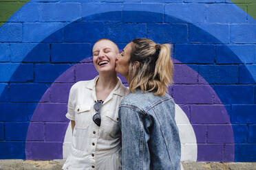 Happy woman kissing on cheeks of non-binary friend in front of colorful wall - ASGF03048