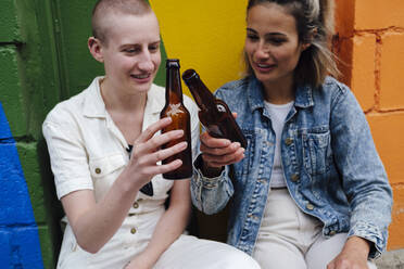 Schwules Paar stößt mit Bierflaschen an, die vor einer bunten Wand stehen - ASGF03049