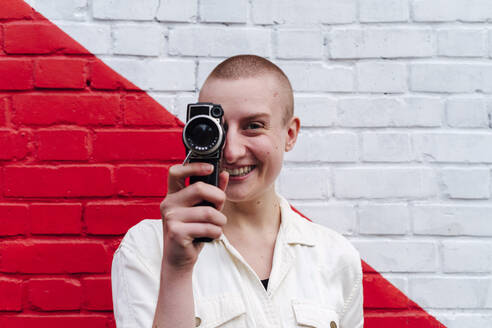Non-binary person covering eye with video camera in front of two toned wall - ASGF03092