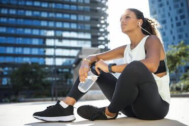 Thoughtful woman sitting with water bottle on footpath - JSRF02293