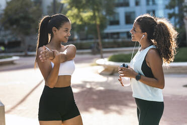 Smiling teenage girl exercising and talking to friend on sunny day - JSRF02303