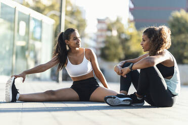 Happy teenage girl exercising and talking to friend sitting on footpath - JSRF02309