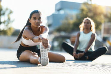 Teenage girl practicing stretching exercise with friend sitting in background on sunny day - JSRF02311