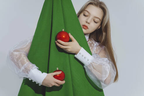 Mädchen hält rote Kugeln und umarmt einen abstrakten Weihnachtsbaum vor weißem Hintergrund - IEF00221