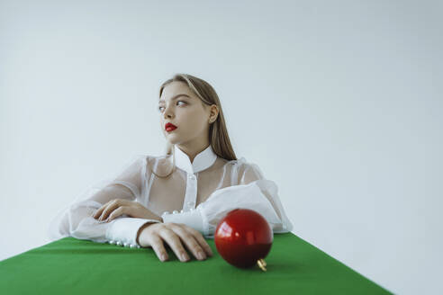 Nachdenkliches Mädchen sitzt mit rotem Weihnachtsschmuck am Tisch vor weißem Hintergrund - IEF00233