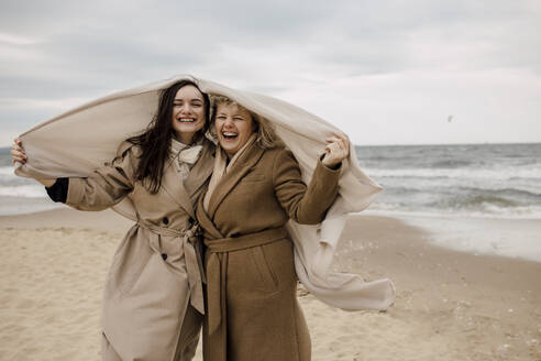 Happy mother and daughter with blanket standing at beach - VIVF00181