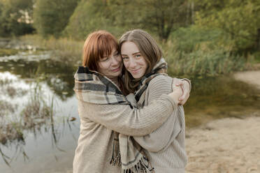 Mother embracing daughter wearing sweater near lake - VIVF00214