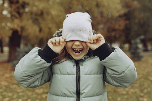 Happy girl covering face with knit hat in autumn park - JBUF00126