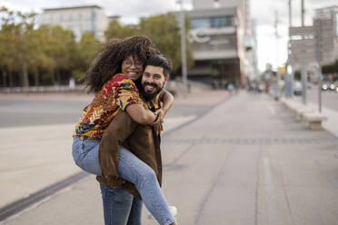 Happy young man giving piggyback ride to girlfriend at footpath - JCCMF07958