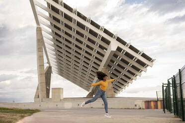 Happy young woman with arms outstretched enjoying in front of solar panels - JCCMF07982