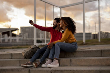 Glückliche junge Frau mit Freund nimmt Selfie durch Smartphone bei Sonnenuntergang - JCCMF07987