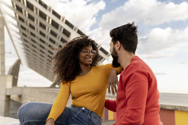 Smiling young woman sitting with boyfriend under cloudy sky - JCCMF08008