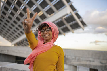 Glückliche junge Frau mit Kopftuch, die vor einer Tafel das Friedenszeichen zeigt - JCCMF08019