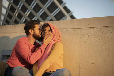 Junger Mann küsst Frau auf die Wange vor einer Mauer sitzend bei Sonnenuntergang - JCCMF08022
