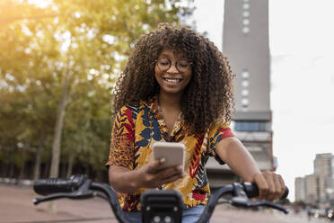 Glückliche junge Frau sitzt auf einem Fahrrad und benutzt ein Smartphone - JCCMF08035