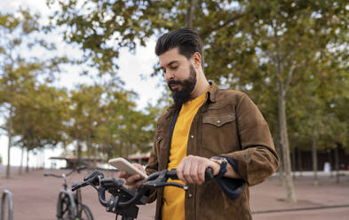 Young man using smart phone standing with bicycle in front of tree - JCCMF08040