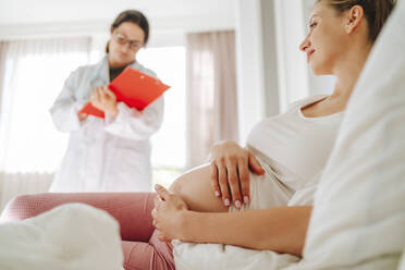 Smiling pregnant woman lying on bed with doctor in background - MDOF00210