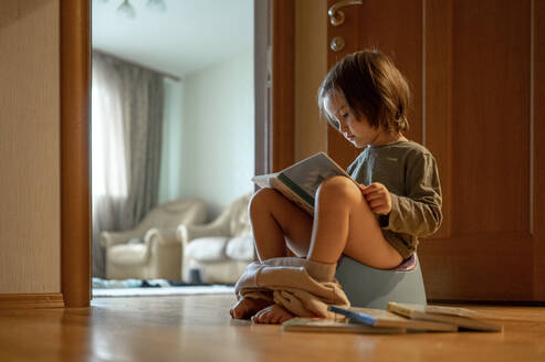 Niedlicher Junge sitzt auf dem Töpfchen und liest ein Buch zu Hause - ANAF00513