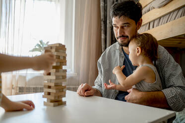 Vater mit Sohn, der ein Spiel zum Entfernen von Blöcken auf dem Tisch zu Hause betrachtet - ANAF00518