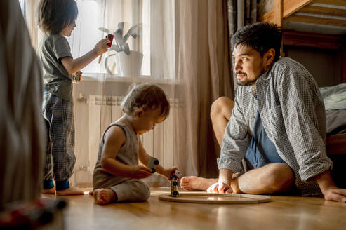 Vater sitzt auf dem Boden und seine Söhne spielen zu Hause - ANAF00525