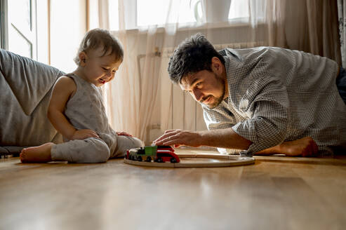 Sohn sieht seinen Vater an, der mit einer Spielzeugeisenbahn auf dem Hausboden spielt - ANAF00529