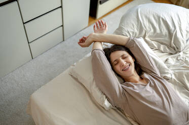 Smiling woman with hands raised waking up in bed at home - ANAF00532