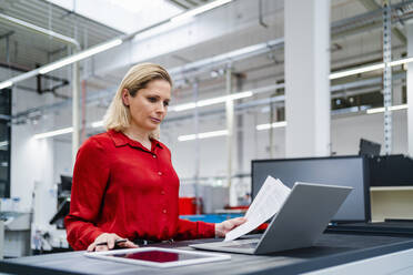 Businesswoman with documents working on laptop in factory - DIGF19417