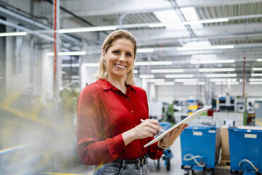 Glückliche Geschäftsfrau mit Tablet-PC in der Fabrik stehend - DIGF19455