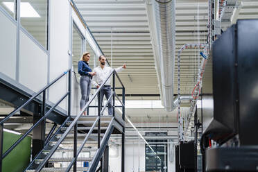 Business colleagues reviewing machinery standing on staircase in factory - DIGF19472