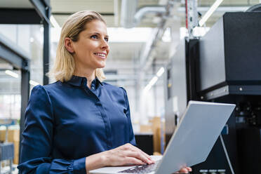 Happy businesswoman wearing blue shirt holding laptop in factory - DIGF19482