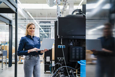 Smiling businesswoman with laptop standing near machinery in industry - DIGF19484