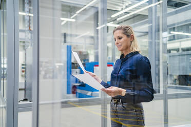 Smiling businesswoman reviewing documents in factory seen through glass - DIGF19500