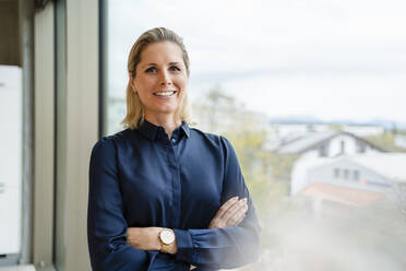 Smiling businesswoman with arms crossed by window in office - DIGF19576