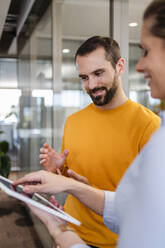 Smiling businessman gesturing by colleague using tablet PC at office - DIGF19606