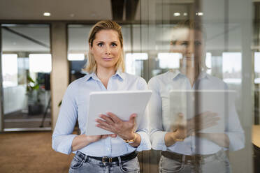 Smiling businesswoman with tablet PC leaning on glass wall at workplace - DIGF19608