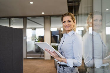 Happy businesswoman with tablet PC leaning on glass wall at office - DIGF19610