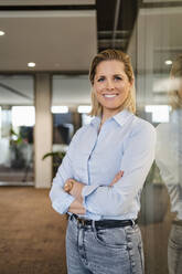 Happy businesswoman with arms crossed leaning on glass wall - DIGF19613