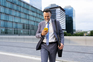 Smiling businessman using smart phone walking on street - OIPF02701